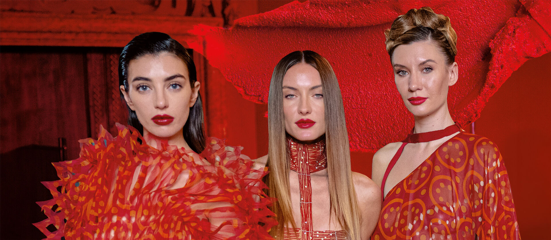 Three models with three different hairstyles and red and orange dresses all done perfectly outlined red lips, a universal symbol of defiance and desire, in front of a smear of red lipstick.