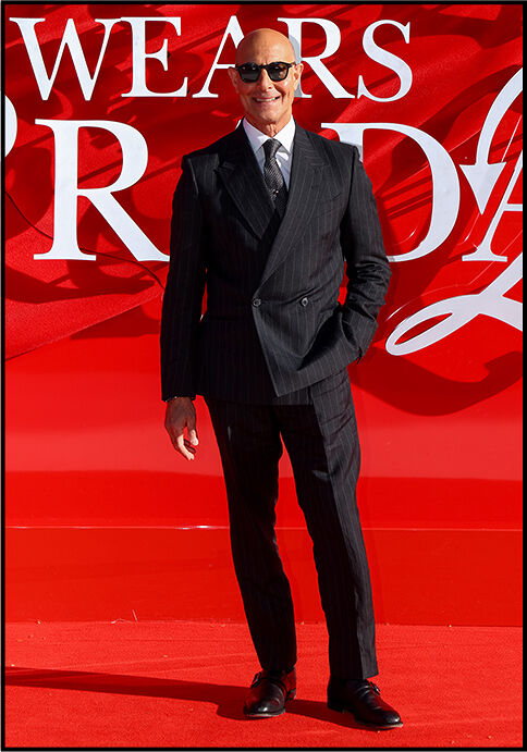 Stanley Tucci wears a classic black suit and sunglasses, standing in front of The Devil Wears Prada 2 logo.