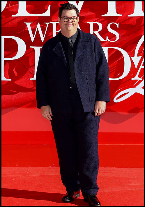 Caleb Hearon smiles softly while wearing a navy suit with a black shirt and tie on the red carpet.