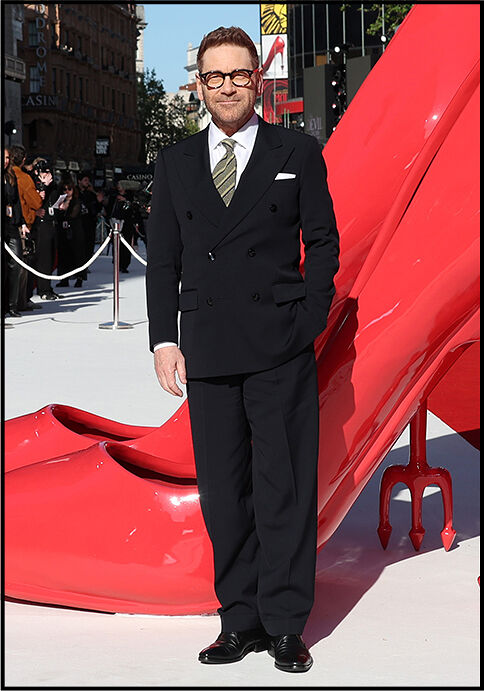 Kenneth Branagh stands in a black suit with a green striped tie in front of the iconic oversized The Devil Wears Prada red heels.