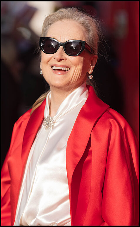 Meryl Streep smiles while wearing oversized sunglasses, a red jacket, and a white silk top with a brooch at the neckline.