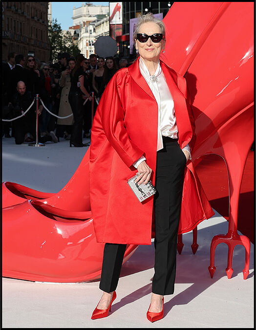 Meryl Streep poses in a white blouse and tailored black pants, layered with a bright red jacket, standing in front of a crowd in London.