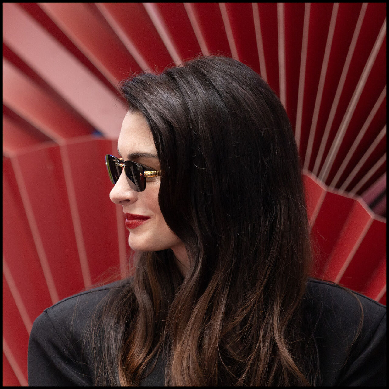 A close up of Anne Hathaway looking off to the side wearing sunglasses and red lipstick.