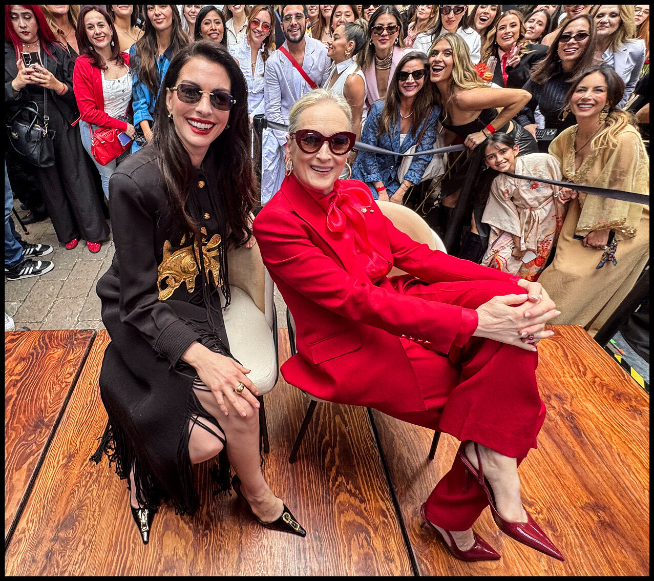 Anne Hathaway and Meryl Streep sitting on chairs with their legs crossed in front of a large crowd.