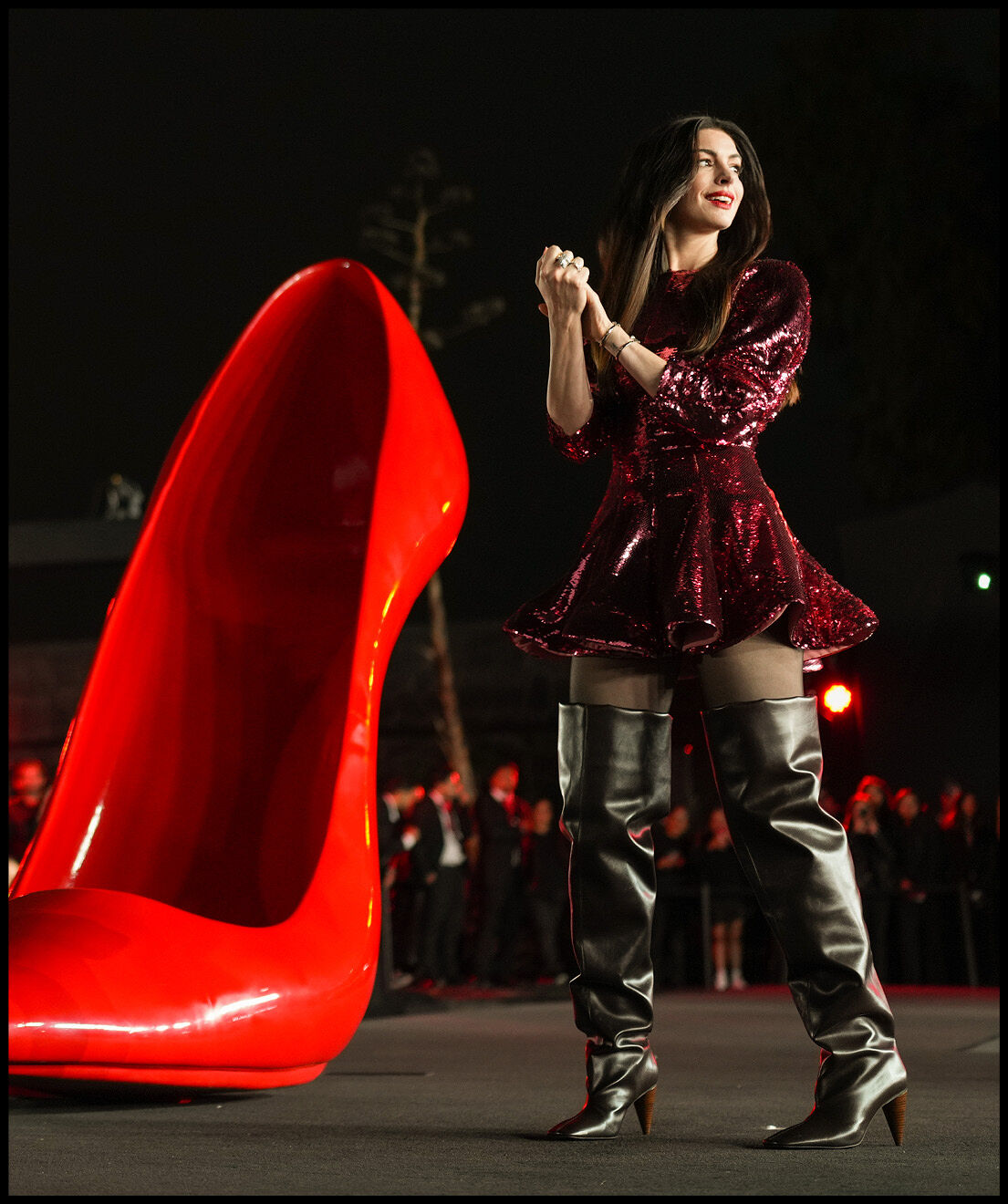 Anne Hathaway with her hands clasped in a sparkly red dress and thigh high black boots next to an oversized red heel. 