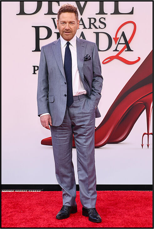 Kenneth Branagh stands in a gray suit with a navy tie, one hand in his pocket.