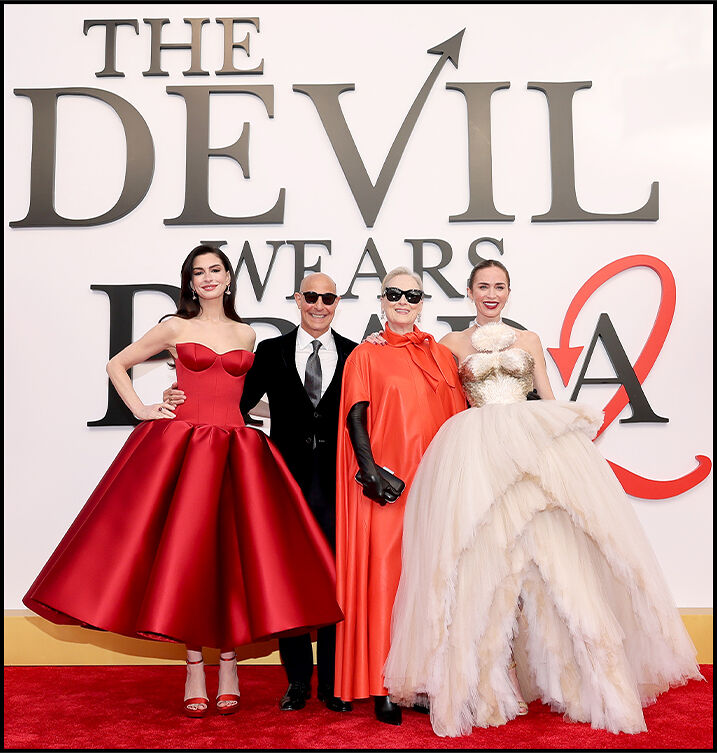 Anne Hathaway, Stanley Tucci, Meryl Streep, and Emily Blunt stand arm in arm, smiling in front of The Devil Wears Prada 2 logo.