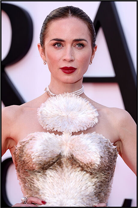 Close‑up of Emily Blunt with a bold red lip and slicked‑back hair, highlighting the feather detailing on the bodice of her dress.