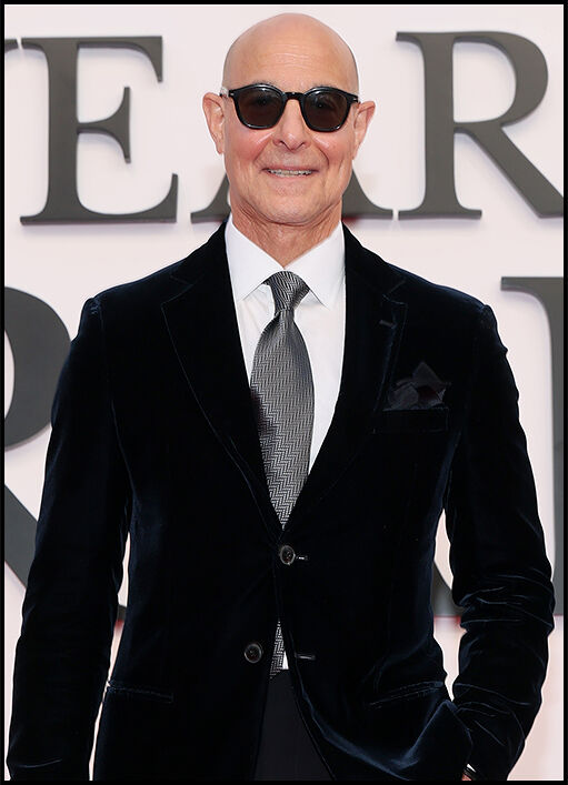 Stanley Tucci smiles at the camera wearing a black suit, gray tie, and black sunglasses.