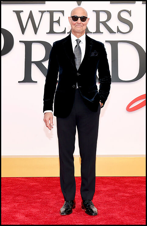 Stanley Tucci stands on the red carpet with one hand in his pocket, dressed in an all‑black suit.