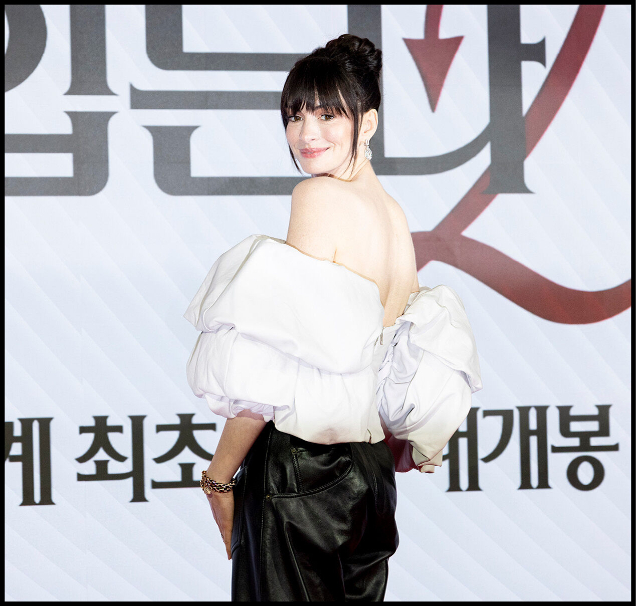 Anne Hathaway smiling over her shoulder wearing a puffy white top and black leather pants.