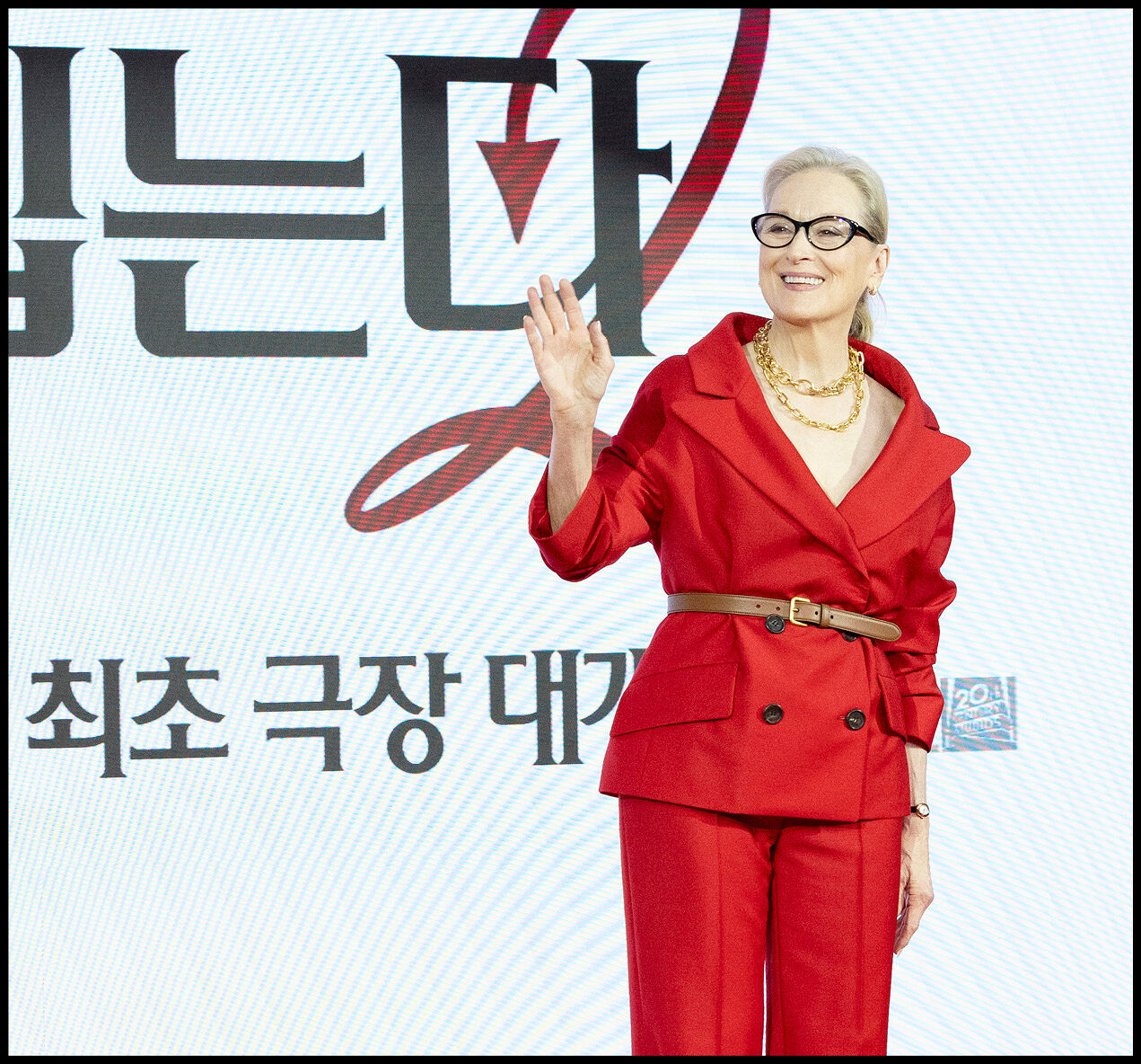 Meryl Streep waving in her red suit in front of the Korean The Devil Wears Prada 2 logo.