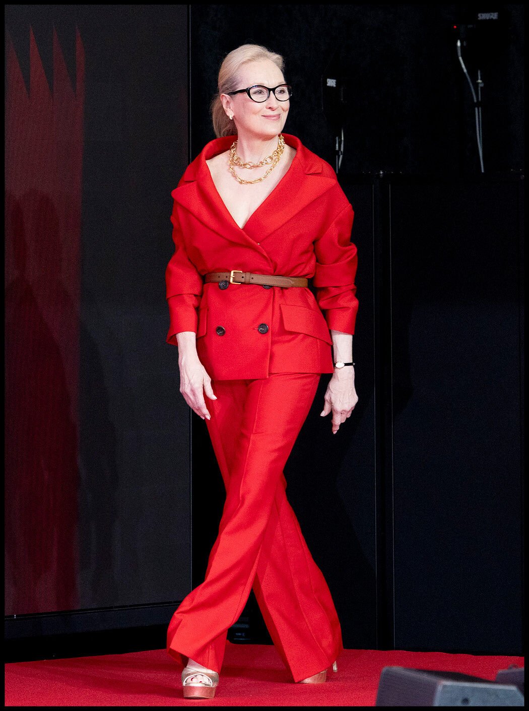 Meryl Streep walking the red carpet in a bold red suit, brown belt, and layered gold necklaces. 