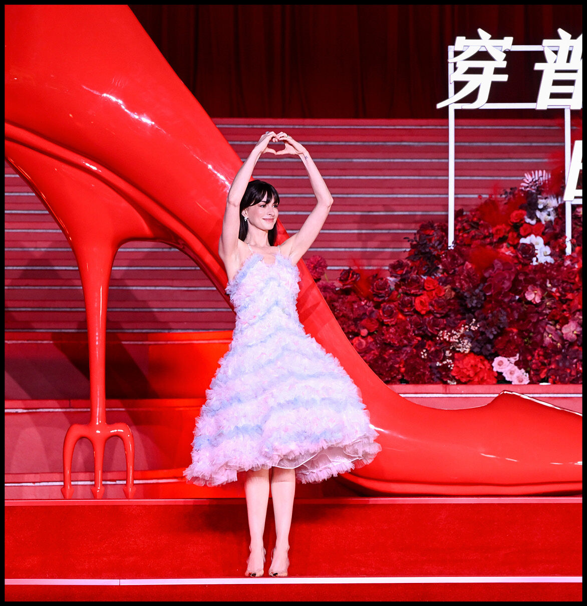 Anne Hathaway in a pastel mid-length dress with her hands above her head in the shape of a heart.