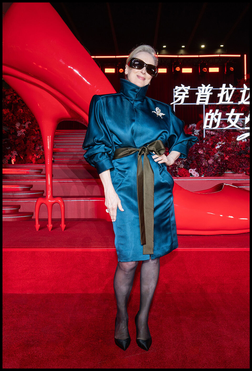 Meryl Streep standing with one hand on her hip in front of an oversized red heel.