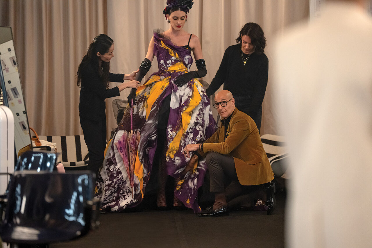 Nigel Kipling, deputy editor of Runway (portrayed by the incomparable Stanley Tucci), makes sure a model’s purple and gold evening gown falls just so minutes before she appears on the catwalk at the magazine’s Milan Fashion Week show. Two assistants also make adjustments to the gloves and ruffles.
