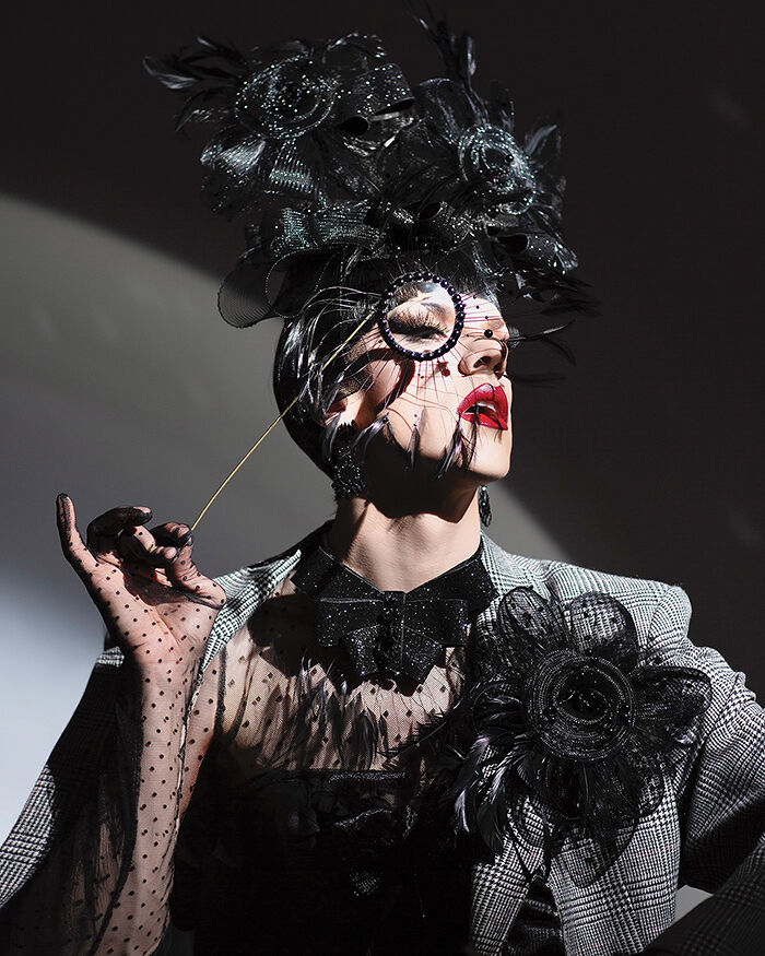 Red-lipped Utica Queen, the drag persona of Ethan Mundt, peeks through a bedazzled and feathered monocle while wearing a tailored blazer, sheer Swiss dot gloves and voluminous headpiece in all black, which he declares the new power color of quiet queer resilience and resistance.
