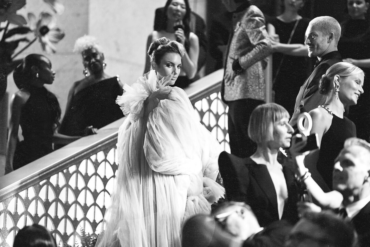A model wearing a flowy dress with a statement neck looks over her shoulder at photographers while on the cerulean carpet at the Runway Gala.