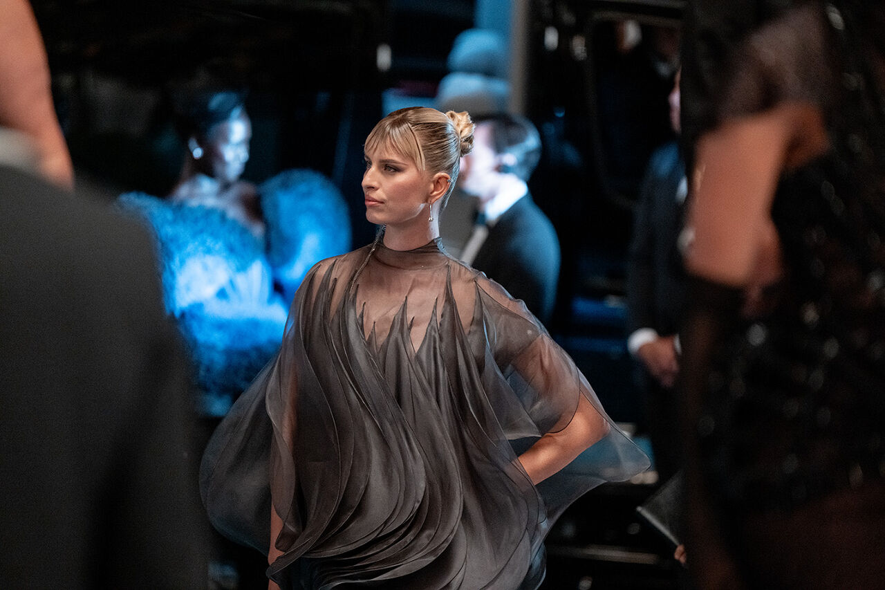 A blonde model with bangs walks with her hand on her hip in a gray sheer wavy gown at the Runway Gala.