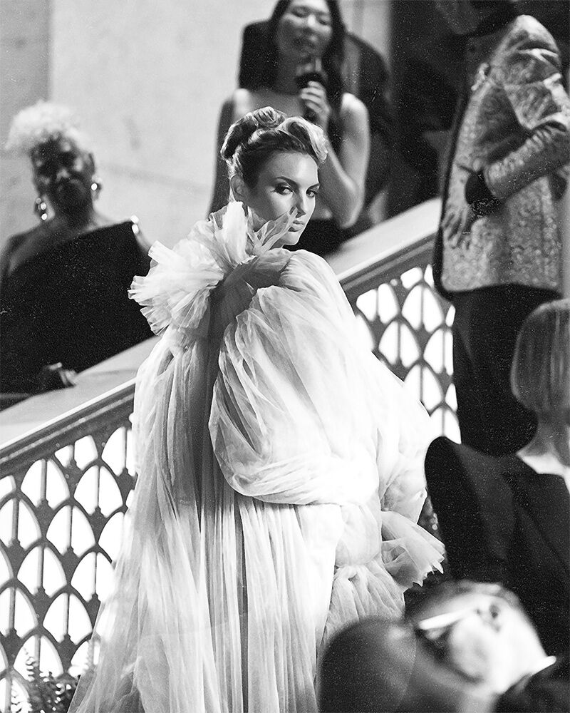 A model wearing a flowy dress with a statement neck looks over her shoulder at photographers while on the cerulean carpet at the Runway Gala.