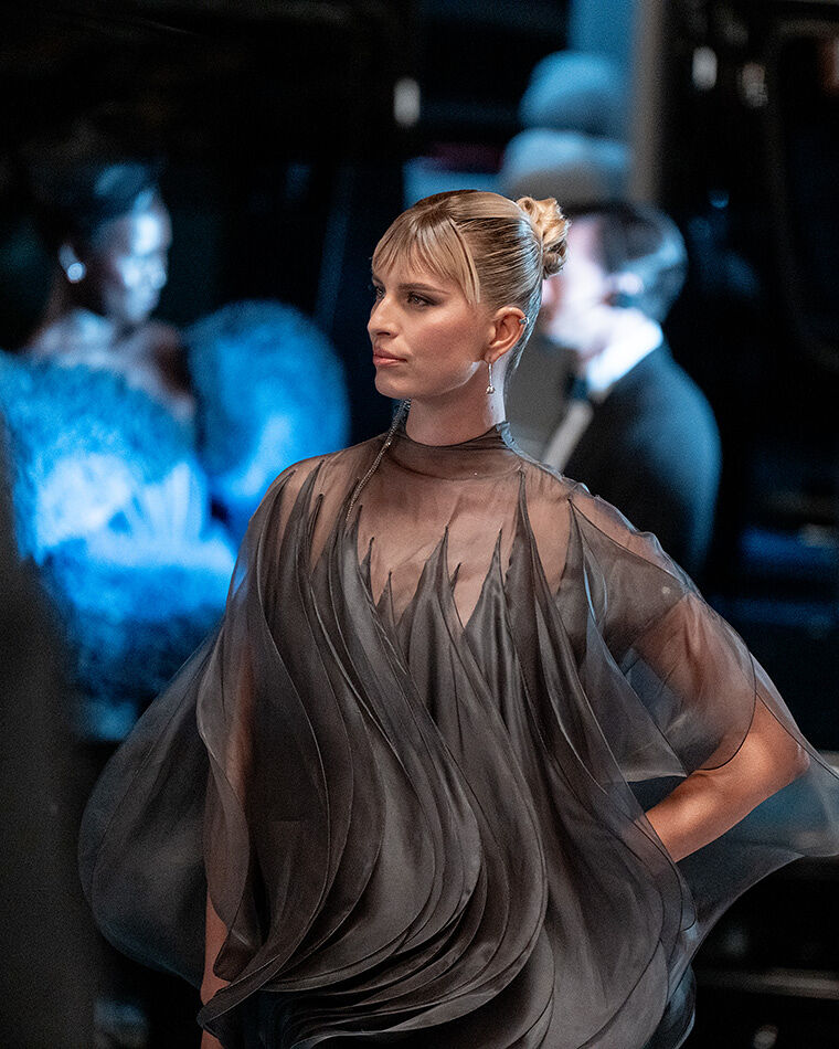 A blonde model with bangs walks with her hand on her hip in a gray sheer wavy gown at the Runway Gala.
