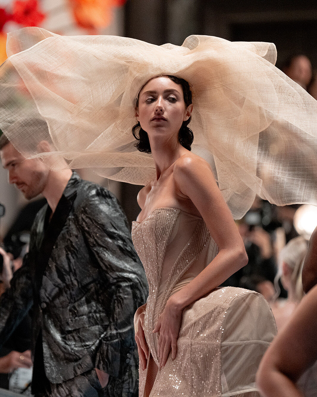 To evoke the Spring Florals theme at this year’s Runway Gala, a model wears a strapless nude bodysuit with hip bustles and details that sparkle like morning dew as well as a giant gauzy headpiece that evokes a blossom.
