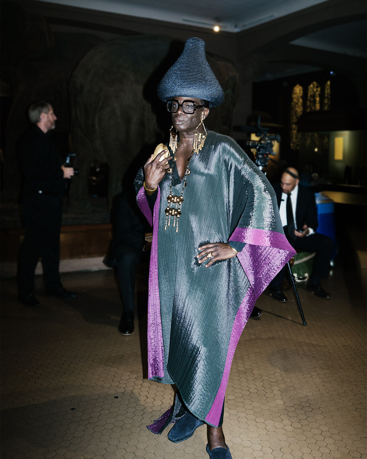 An attendee of the Runway Gala wears a shiny high-fashion caftan, lots of bold gold jewelry, strong square black frames, blue suede shoes and a woven basket hat shaped like a Hershey Kiss.