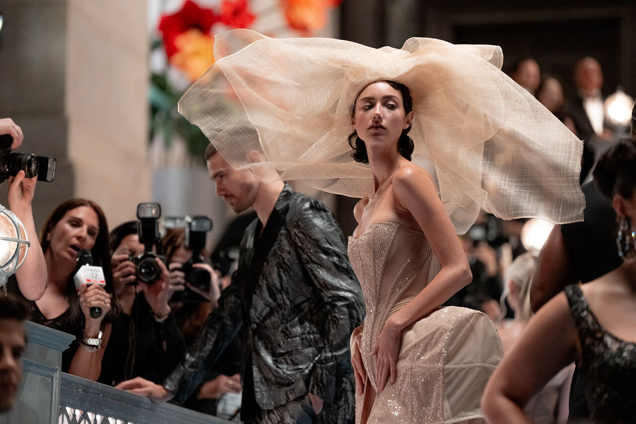 To evoke the Spring Florals theme at this year’s Runway Gala, a model wears a strapless nude bodysuit with hip bustles and details that sparkle like morning dew as well as a giant gauzy headpiece that evokes a blossom.