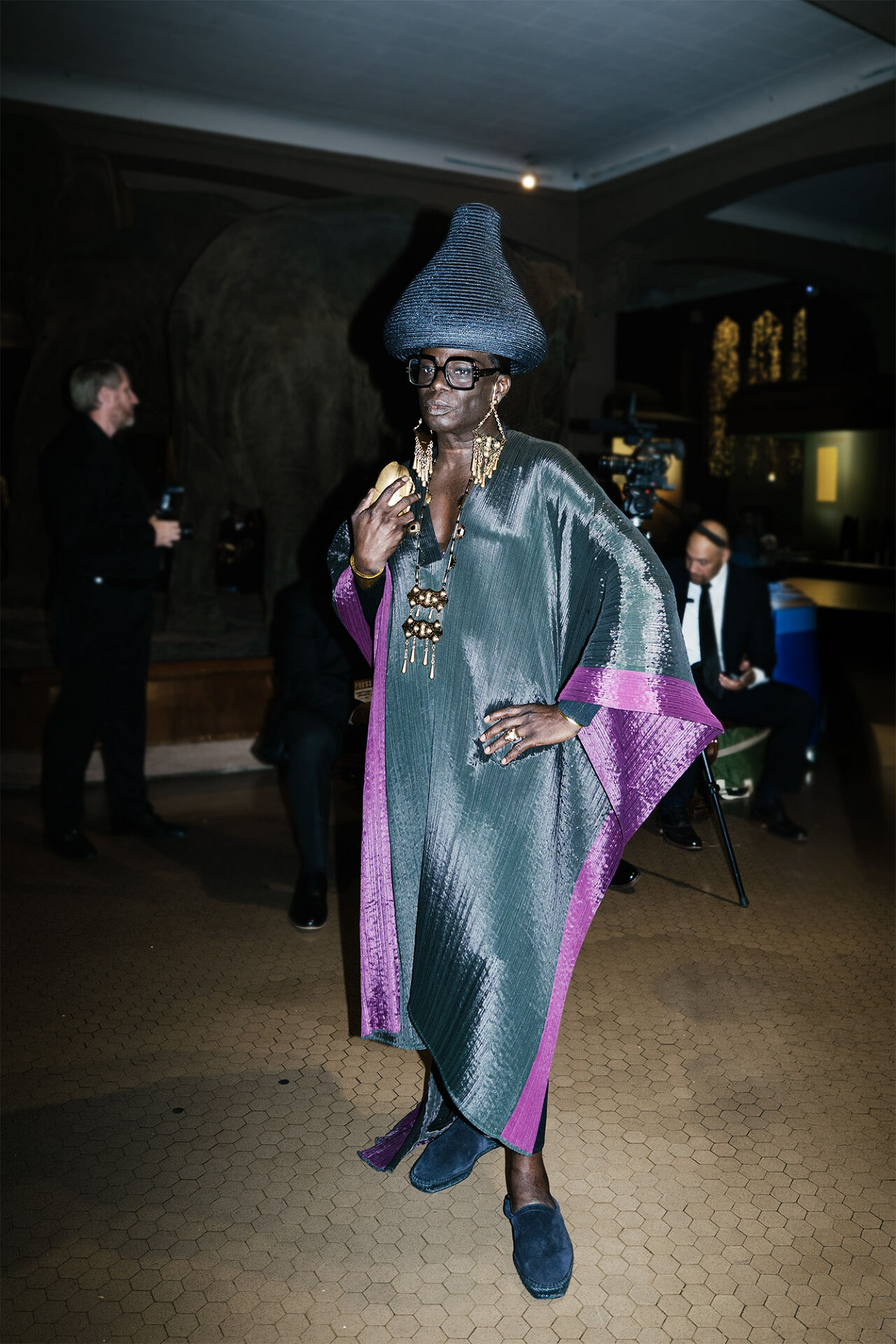 An attendee of the Runway Gala wears a shiny high-fashion caftan, lots of bold gold jewelry, strong square black frames, blue suede shoes and a woven basket hat shaped like a Hershey Kiss.