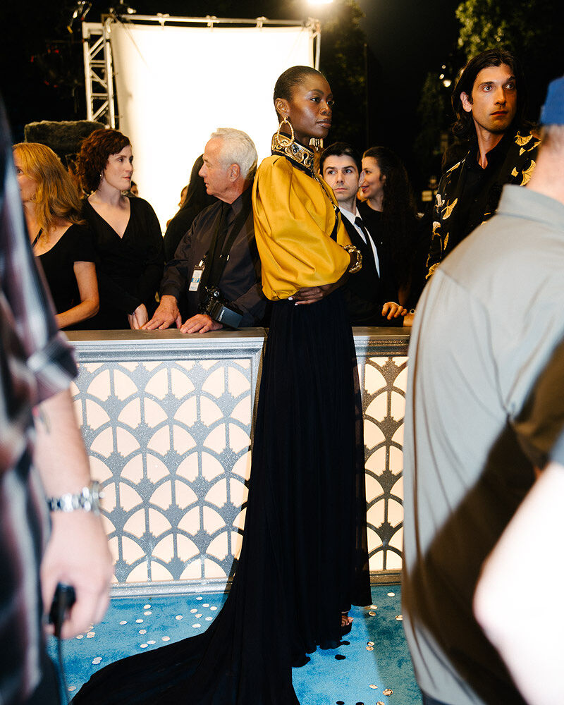 A gorgeous guest in a gold blouse and elegant black skirt with a train stands on a confetti-covered cerulean carpet at the annual Runway Gala. 