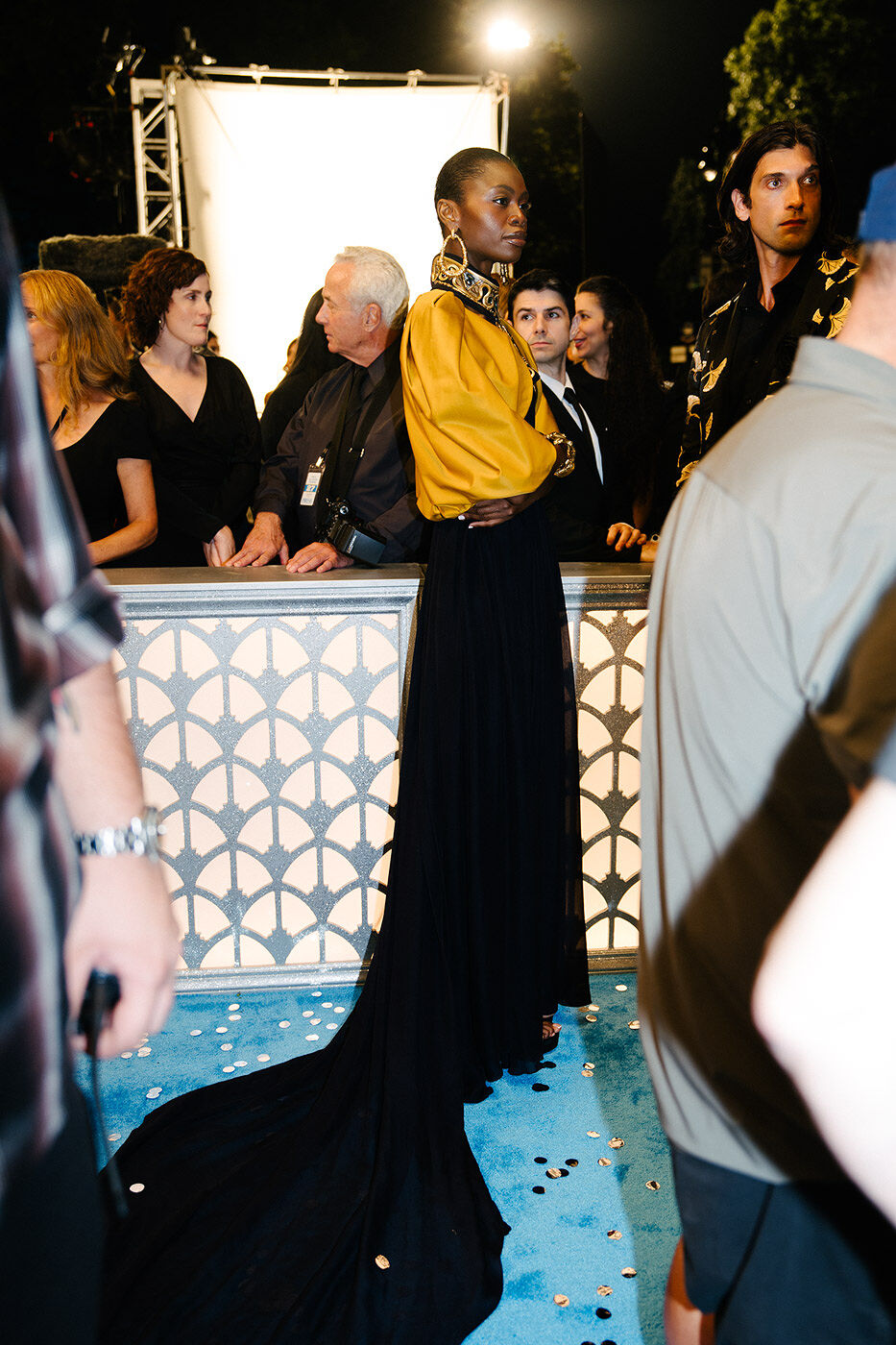 A gorgeous guest in a gold blouse and elegant black skirt with a train stands on a confetti-covered cerulean carpet at the annual Runway Gala. 