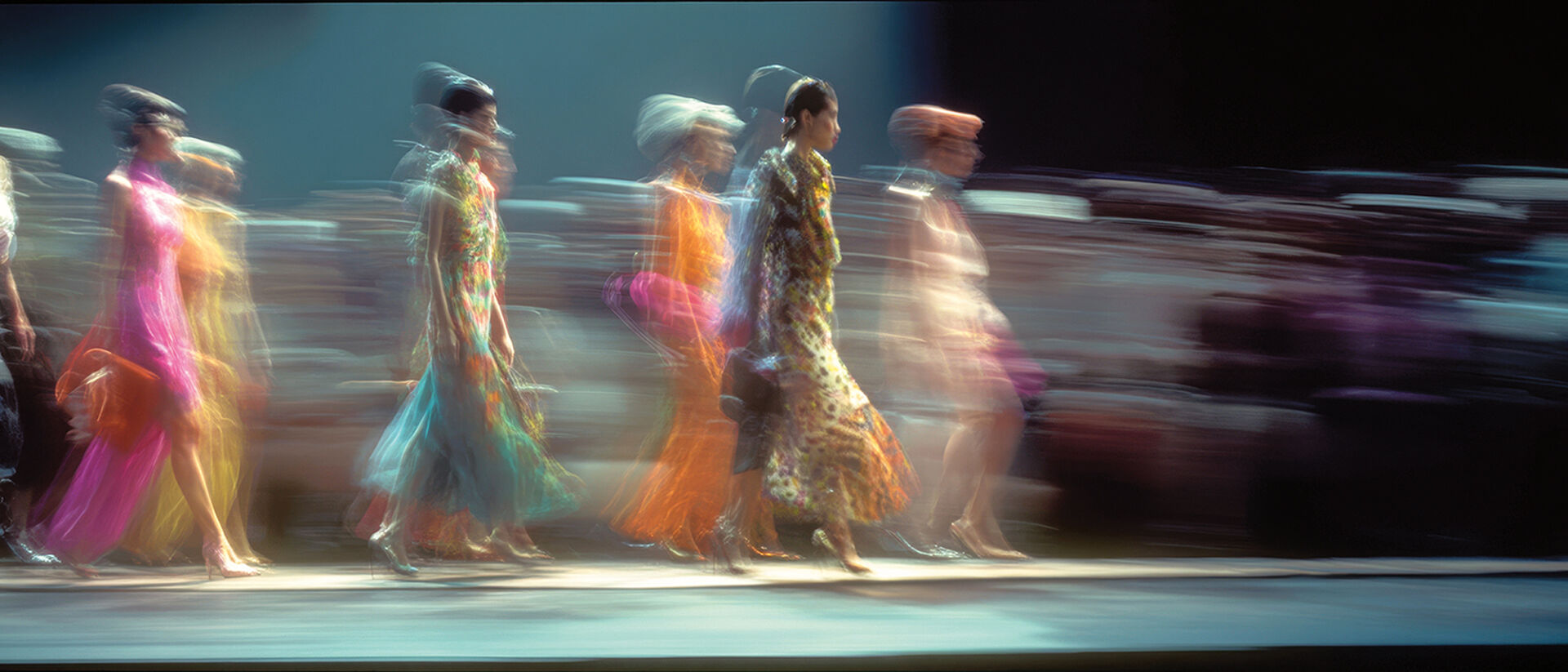 Group of models in vibrantly colored dresses walking the catwalk together in the finale of a runway show.