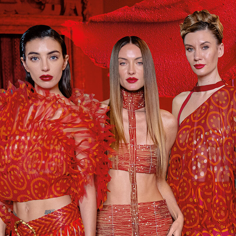 Three models with three different hairstyles and red and orange dresses all don perfectly outlined red lips, a universal symbol of defiance and desire, in front of a smear of red lipstick and a scarlet background.