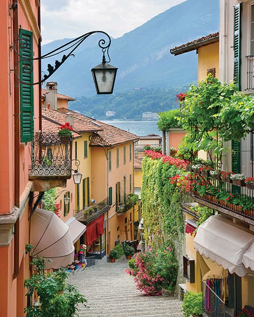 A picturesque sloped side street in one of Lake Como’s many quaint hillside villages features flowers, dripping vines, wrought iron balconies, colorful shutters, hanging wall scones and stone stairs.