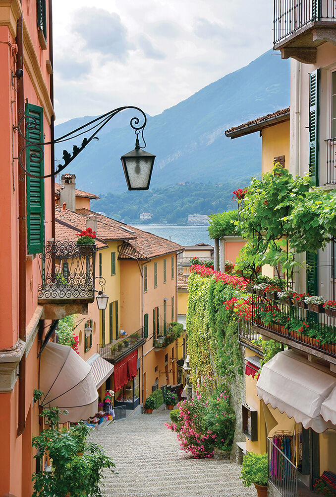 A picturesque sloped side street in one of Lake Como’s many quaint hillside villages features flowers, dripping vines, wrought iron balconies, colorful shutters, hanging wall scones and stone stairs.